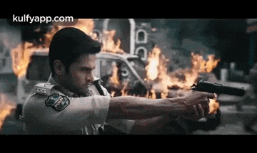 a man is holding a gun in front of a burning car .
