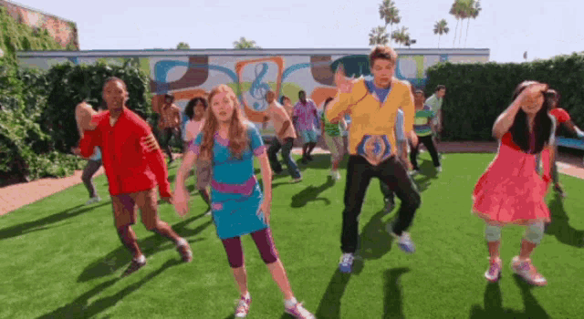 a group of people are dancing on a grassy field in front of a wall with a treble clef on it