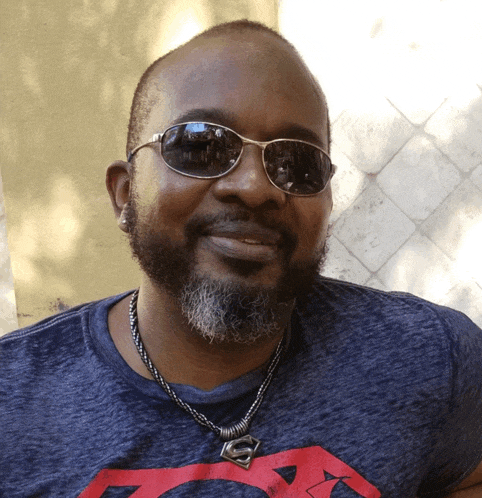 a man wearing sunglasses and a superman necklace smiles for the camera