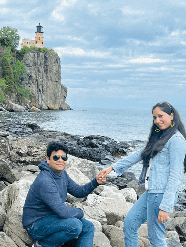 a man and a woman are holding hands on a rocky shoreline with a lighthouse in the background