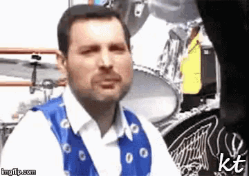 a man wearing a blue vest and a white shirt is sitting in front of a drum set .
