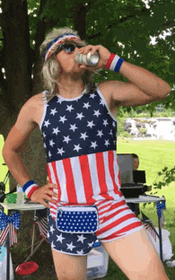 a man wearing an american flag tank top and shorts drinks from a can