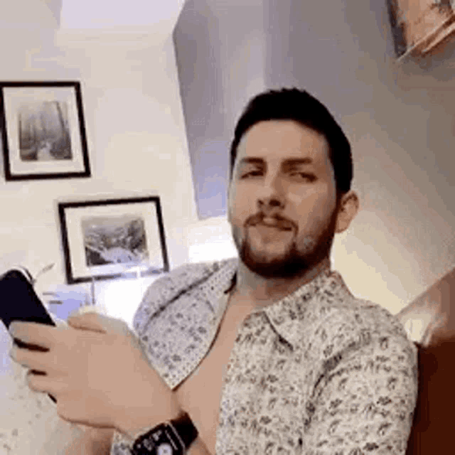 a shirtless man with a beard is sitting on a bed holding a cell phone .