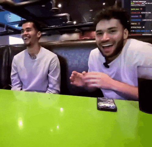two men are sitting at a table with a green table cloth and a remote control on it