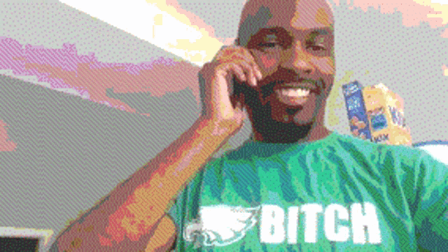 a man wearing a green shirt that says " bitch " is talking on a cell phone
