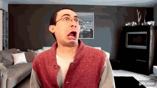 a man wearing glasses and a red vest is making a funny face in a living room ..