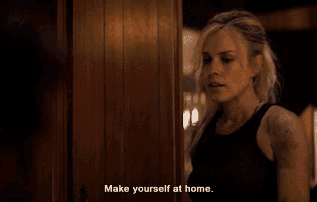 a woman in a black tank top is standing in front of a wooden door and says make yourself at home
