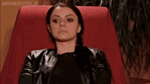 a woman in a leather jacket is sitting in a red chair and making a funny face .