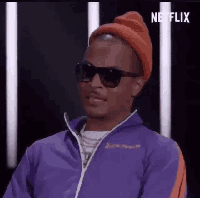 a man wearing a purple jacket , sunglasses , and an orange hat is sitting in front of a netflix sign .