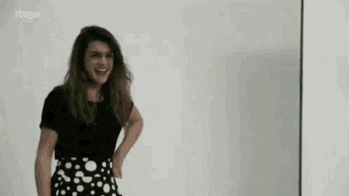 a woman in a black shirt and polka dot skirt is standing in front of a white wall and smiling .