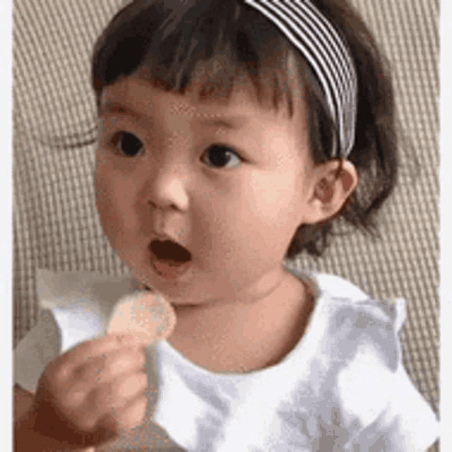 a baby girl wearing a headband is eating a piece of food with her mouth open .