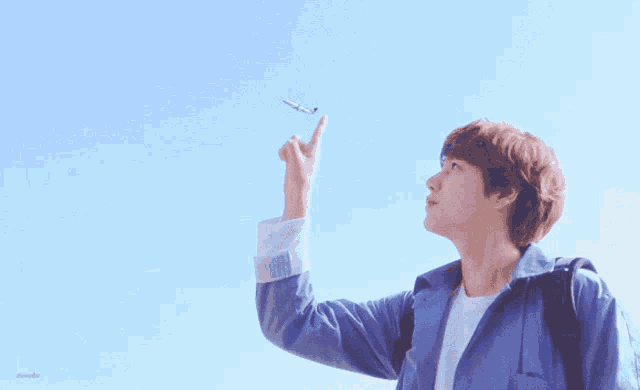 a young man in a blue jacket is pointing up at a small airplane in the sky