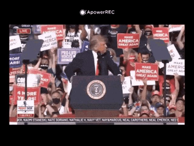 a man stands at a podium in front of a crowd holding a sign that says make america great again
