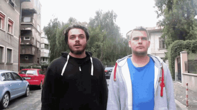 two men standing next to each other with one wearing a blue shirt and the other a black hoodie