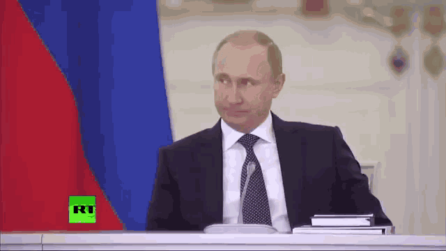 a man in a suit and tie is sitting at a desk in front of a flag with rt on the screen behind him