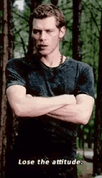 a man in a black shirt is standing in the woods with his arms crossed and says lose the attitude .