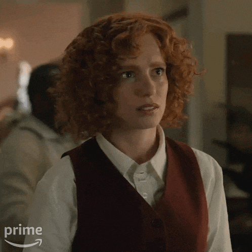 a woman with red curly hair is wearing a vest and white shirt .