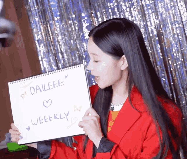 a woman in a red jacket is holding a notepad with daieeee weekly written on it