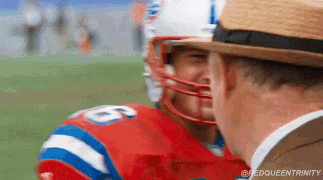 a football player wearing a helmet is talking to a man wearing a hat on a field .