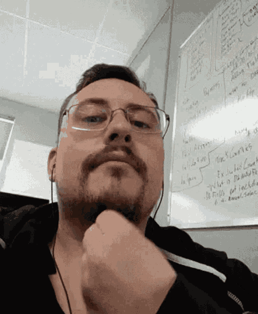 a man with glasses and a beard looks at the camera in front of a whiteboard that says ' a '