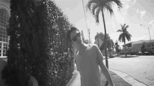a man wearing sunglasses is walking down a sidewalk in front of a palm tree