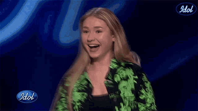 a girl in a green jacket is singing into a microphone in front of an idol logo .
