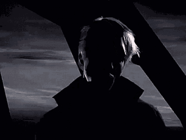 a man in a black coat is standing in the dark looking out a window .