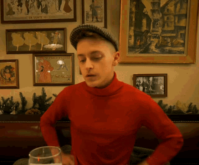 a man in a red turtleneck sweater stands in front of a wall of framed pictures one of which says en vente