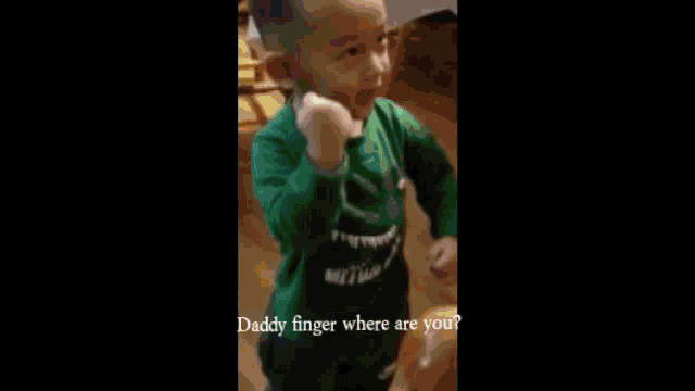 a little boy in a blue shirt is standing in front of a table and giving the middle finger .