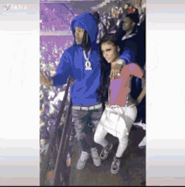 a man in a blue hoodie and a woman in a pink shirt are standing next to each other in front of a crowd .