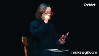 a woman wearing glasses and a black sweater is sitting in a chair holding a book and a magazine .
