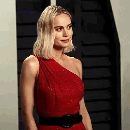 a woman in a red dress with a black belt is standing in front of a staircase .