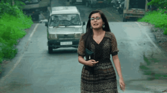 a woman is walking down a road with a car behind her that has a license plate that starts with a letter a