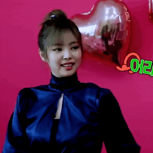 a woman in a blue shirt is standing in front of a pink wall with a heart shaped balloon behind her .