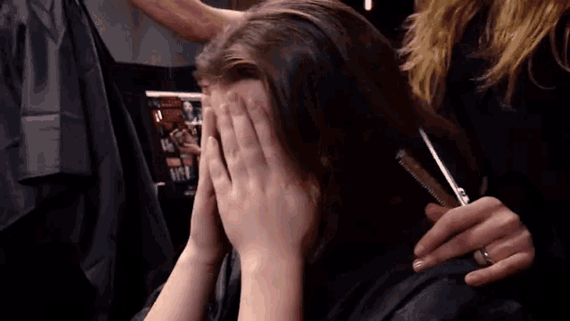a woman is covering her face with her hands while a hairdresser brushes her hair