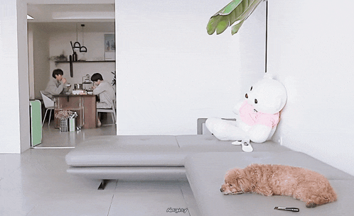 a teddy bear sits on a couch in a living room with two men sitting at a table