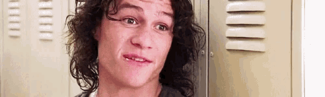 a man with long curly hair is standing in front of a locker making a funny face .