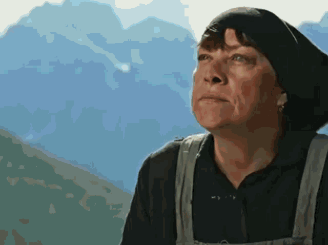 a woman wearing a black hat and overalls looks up at the sky with mountains in the background