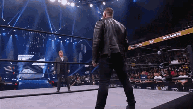 a man in a suit stands in a wrestling ring with a banner that says dynamite on it