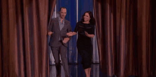 a man in a suit and a woman in a black dress are walking through a curtain on a stage .