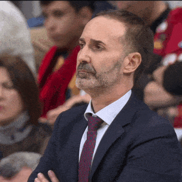 a man in a suit and tie sits in a crowd with his arms crossed