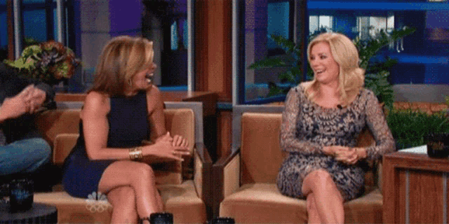 two women are sitting on a couch laughing and one of them is wearing a blue dress