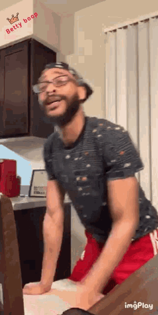 a man wearing glasses and a hat is dancing in a kitchen with the words betty boop on the wall behind him .