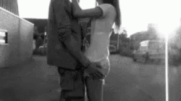 a black and white photo of a man and a woman kissing in front of a building .
