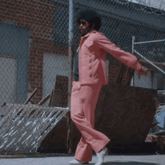 a man in pink pants is standing in front of a chain link fence and a brick building