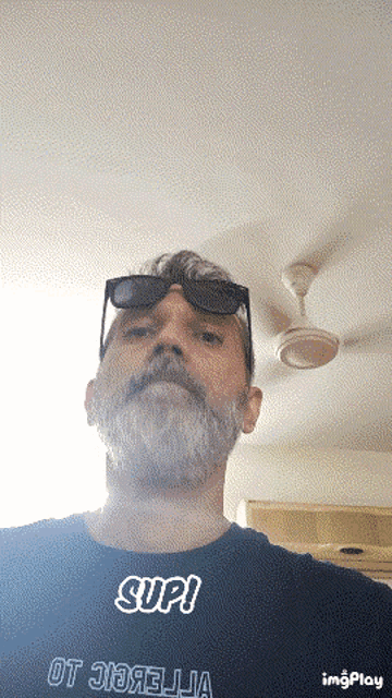 a man with a beard wears sunglasses and a shirt that says sup