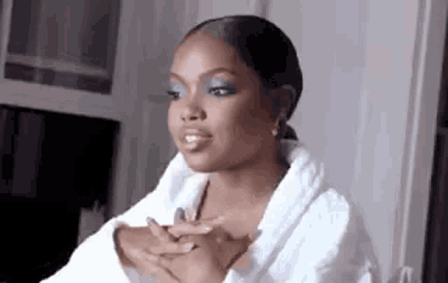 a woman wearing a white robe and blue eyeshadow is sitting in a room with her hands folded .