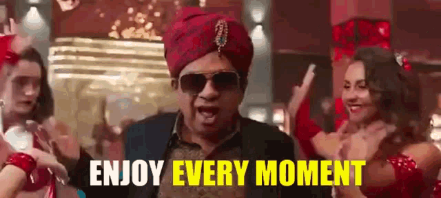 a man wearing a turban and sunglasses says enjoy every moment while dancing with women .