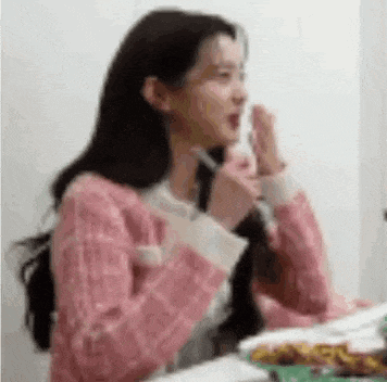 a woman in a pink sweater is sitting at a table with a plate of food and a pen in her hand .