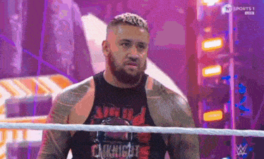 a man with a beard is standing in a wrestling ring wearing a black tank top .